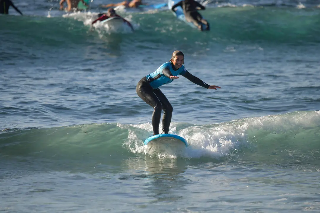 Alumna de pie sobre la tabla cogiendo una ola durante una clase de surf en Playa de Las Américas